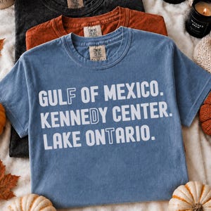Op de afbeelding: Een blauw t-shirt met korte mouwen en witte tekst die GULF OF MEXICO, KENNEDY CENTER, LAKE ONTARIO leest. Het shirt heeft een Comfort Colors merklabel bij de binnenhalslijn en ligt over een roestkleurig en een zwart t-shirt heen.