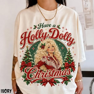May include: An ivory t-shirt featuring a graphic print of a blonde woman in a red fringed outfit, surrounded by two Christmas trees, red bows, and stars. The text reads Have a Holly Dolly Christmas in red and green script and block lettering.