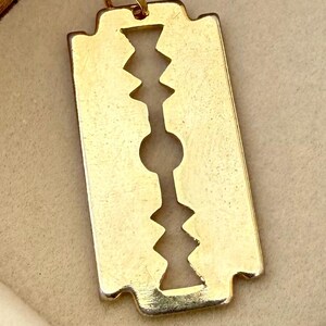 May include: A gold-toned pendant shaped like a classic double-edged razor blade. The pendant features a central cutout design consisting of a circular hole flanked by two jagged, geometric patterns. A small circular jump ring is attached at the top.