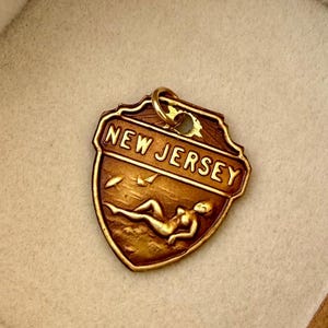 May include: A gold-toned metal pendant shaped like a shield with a jump ring at the top. The pendant features the embossed text NEW JERSEY above a relief scene of a figure reclining on a beach with a small umbrella and sailboat in the background.