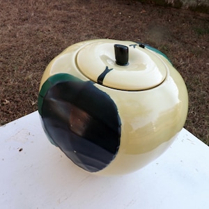 May include: A ceramic cookie jar shaped like a large apple with a glossy cream-colored finish, featuring a black stem handle on the lid and a prominent black and dark green painted patch on the side, creating a distinct contrast against the light body.