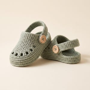 Op de afbeelding: Een paar saliegroene gehaakte babyschoentjes met een ronde neus, een reeks kleine uitsparingen aan de voorkant en een beweegbare hielband die aan elke kant is bevestigd met een enkele lichtgekleurde houten knoop.