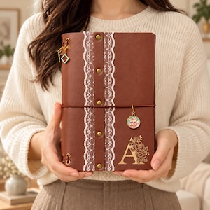 May include: A brown leather journal featuring a vertical white lace strip secured by four brass-colored studs. The cover displays a gold letter A with floral accents, a dangling square charm, and a circular charm with a flower illustration.