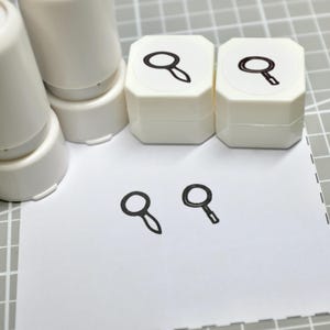 May include: Two white plastic craft stamps with square bases, each featuring a black magnifying glass icon on top. The stamps are positioned above two black ink impressions of magnifying glasses stamped onto a white sheet of paper.