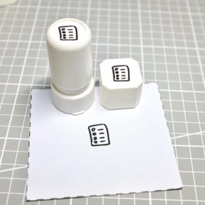 Può includere: Due timbri in plastica bianca con un'icona a forma di lista di controllo nera stampata sulla parte superiore di ciascuno. Un timbro è cilindrico e l'altro è quadrato. È visibile un foglio di carta quadrato bianco con la stessa icona.