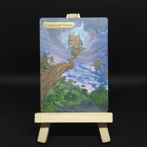 May include: A rectangular trading card titled Command Tower resting on a small wooden easel. The card features a painted illustration of a floating structure above a tree branch, set against a blue and purple sky with green foliage below.