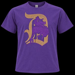 May include: A purple short-sleeve t-shirt featuring a large, stylized letter D graphic on the chest. The letter is outlined in gold-colored rhinestones and filled with purple rhinestones, forming a decorative design across the front of the garment.