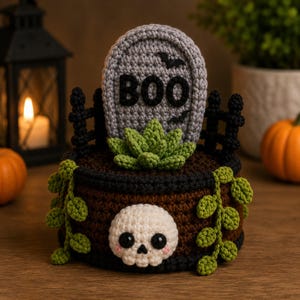 May include: A crocheted Halloween-themed planter featuring a brown base with a white skull, a grey tombstone with the text BOO, a green succulent, and trailing green vines. The design includes a black fence border and small black bats.