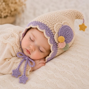 Puede incluir: Un gorro de ganchillo color crema con punta y borde festoneado morado. El gorro presenta una flor morada grande, pequeños pompones amarillos y rosas, y una estrella amarilla en la punta. Cuelgan lazos de ganchillo morados con detalles de estrellas.
