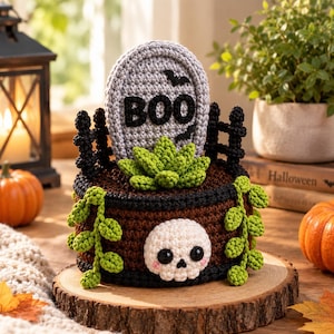 May include: A crocheted Halloween-themed planter featuring a brown base with a white skull, a gray tombstone with the black text BOO, a green succulent, and trailing green vines. The structure is accented with a black fence and black trim.