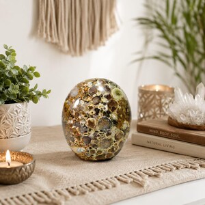 Puede incluir: Una piedra decorativa lisa en forma de huevo con un patrón complejo y moteado de inclusiones circulares en tonos marrón tierra, verde oliva y crema. La superficie tiene un acabado pulido y brillante que refleja la luz sobre su forma.