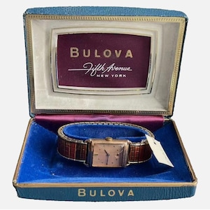 May include: A vintage Bulova Fifth Avenue New York wristwatch with a rectangular gold-tone case and a textured brown expansion band, presented in a blue velvet-lined display box with the brand name printed in gold on the interior and exterior.