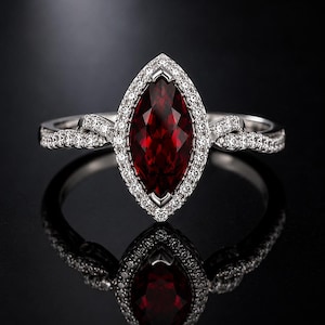 May include: A silver-toned ring featuring a large, marquise-cut deep red gemstone at the center. The stone is surrounded by a halo of small, clear, sparkling stones. The band has a twisted design encrusted with additional small clear stones.