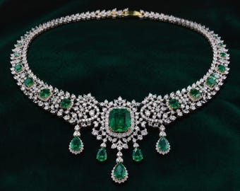 May include: A silver-toned necklace featuring a large emerald-cut green gemstone at the center, surrounded by numerous small clear crystals. The design includes multiple pear-shaped green gemstone drops and a band of clear crystals throughout.