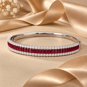 May include: A silver-toned hinged bangle bracelet featuring a central row of rectangular-cut deep red rubies flanked by two rows of small, round, clear white stones. The bracelet has a secure metal clasp mechanism on the side.