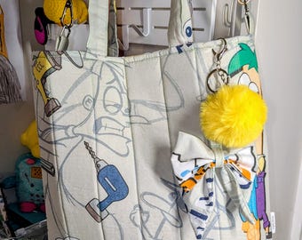 May include: A quilted tote bag featuring a light-colored fabric with a repeating cartoon print of Phineas and Ferb characters and tools. The bag includes two top handles, a detachable black shoulder strap, a fabric bow, and a bright yellow pom-pom.