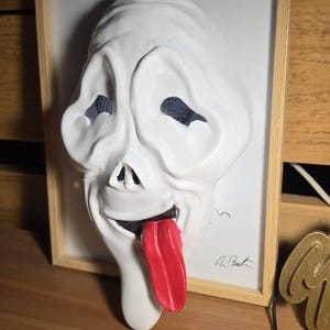 Könnte beinhalten: Eine weiße, dreidimensionale Wandmaske mit einem verzerrten, länglichen Gesicht, hohlen schwarzen Augenhöhlen, einer kleinen Nase und einem weit geöffneten Mund, aus dem eine auffällige, leuchtend rote Zunge herausragt, gerahmt in hellem Holz.