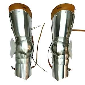May include: A pair of polished steel leg armor greaves featuring articulated knee caps with side wings, connected to upper thigh plates with tan leather straps and buckles for secure attachment, showcasing a metallic silver finish throughout.