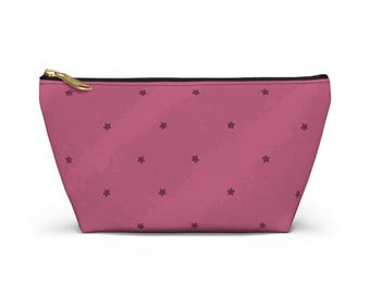 May include: A trapezoidal zippered pouch in a dusty rose pink color featuring a repeating pattern of small, dark maroon five-pointed stars across the exterior surface. The pouch includes a gold-toned metal zipper pull at the top edge.