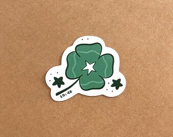 May include: A die-cut sticker featuring a four-leaf clover in shades of green with a white star at its center. The design includes two dark green stars on the sides, small dots, and the text 10:42 printed in black near the clover stem.