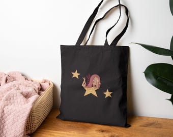 May include: A black tote bag featuring a graphic illustration of a character with long pink hair lying on a large, tan, five-pointed star. Two smaller tan stars are positioned above and to the right of the central figure on the fabric surface.