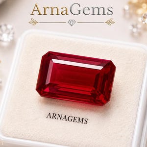 May include: A rectangular, faceted deep red gemstone sits centred in a white square display box. The text ARNAGEMS is printed on the box surface. Above the box, the logo AG ArnaGems is displayed with a small diamond icon underneath.