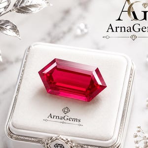 Puede incluir: Una gema roja de corte hexagonal alargado descansa sobre un joyero forrado de terciopelo blanco. La caja presenta el logotipo de ArnaGems y un cierre plateado decorativo. La escena está decorada con hojas plateadas y pequeños cristales.