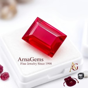 Op de afbeelding: Een levendige, rechthoekig geslepen rode edelsteen op een wit displaydoosje. Het doosje bevat de tekst ArnaGems Fine Jewelry Since 1998 en een goudkleurig AG-logo. Daarnaast liggen twee kleine losse rode stenen, een rood fluwelen zakje en een juweliersloep.