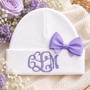 May include: A white beanie featuring a folded brim with a purple embroidered monogram of the letters CLM. A small, purple grosgrain ribbon bow is attached to the right side of the beanie, positioned just above the brim for a decorative accent.
