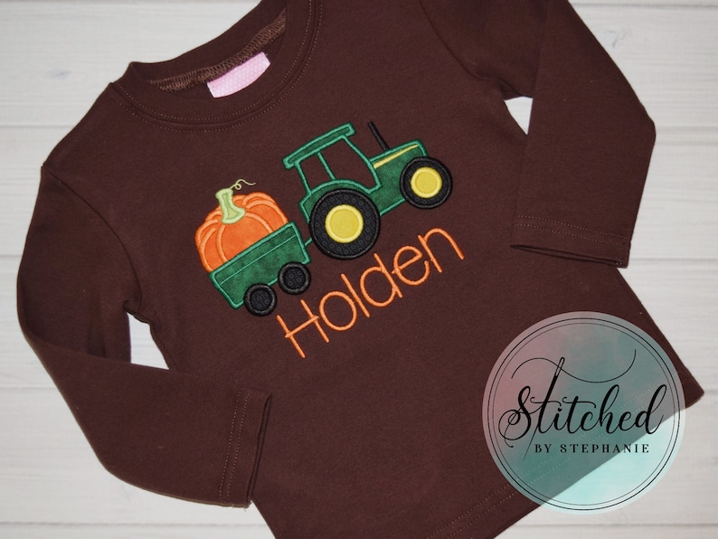 May include: A long-sleeved brown shirt featuring an embroidered design of a green tractor pulling a green wagon with an orange pumpkin. The name "Holden" is embroidered in orange. The shirt is a fall-themed item.