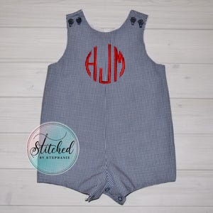 Peut inclure: Barboteuse à carreaux bleus et blancs avec monogramme rouge. La barboteuse a une encolure ronde, des boutons aux épaules et aux jambes, et un monogramme rouge brodé dans un cercle. Le texte "Stitched by Stephanie" est en bas à gauche.