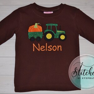 May include: A long-sleeved brown shirt featuring an embroidered design of a green tractor pulling a wagon with an orange pumpkin. The name "Nelson" is embroidered in orange. The shirt is a children's apparel item.