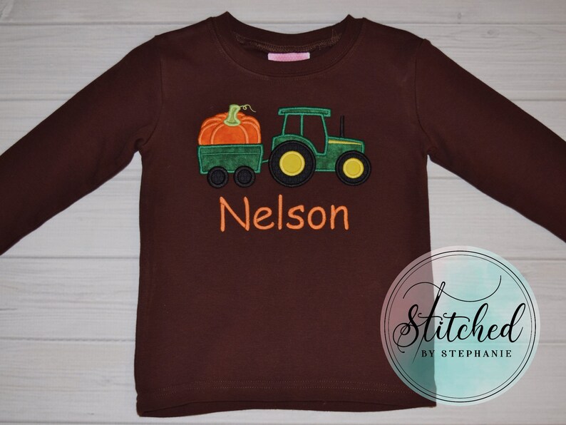 May include: A long-sleeved brown shirt featuring an embroidered design of a green tractor pulling a wagon with an orange pumpkin. The name "Nelson" is embroidered in orange. The shirt is a children's apparel item.