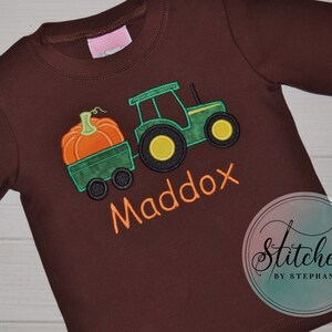 May include: A long-sleeved brown shirt featuring an embroidered design of a green tractor pulling a wagon with an orange pumpkin. The name "Maddox" is embroidered in orange. The shirt has a round neckline.