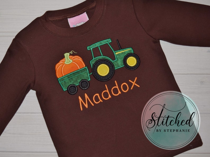May include: A long-sleeved brown shirt featuring an embroidered design of a green tractor pulling a wagon with an orange pumpkin. The name "Maddox" is embroidered in orange. The shirt has a round neckline.
