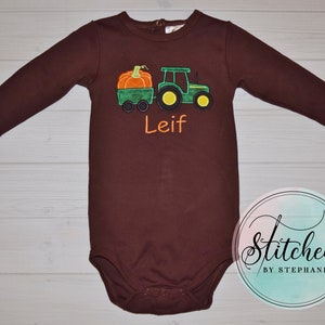 May include: A brown long-sleeved baby bodysuit featuring an embroidered green tractor pulling a green wagon with an orange pumpkin. The name "Leif" is embroidered in orange. The design is on a brown bodysuit.