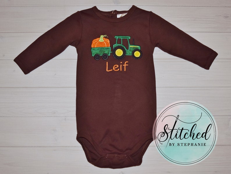 May include: A brown long-sleeved baby bodysuit featuring an embroidered green tractor pulling a green wagon with an orange pumpkin. The name "Leif" is embroidered in orange. The design is on a brown bodysuit.