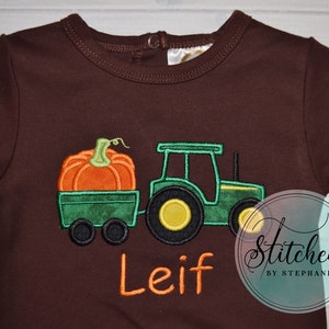 May include: Brown long-sleeve baby bodysuit with an embroidered design. The design features a green tractor pulling a wagon with an orange pumpkin. The name "Leif" is embroidered in orange. The text "Stitched by Stephanie" is in a circle.