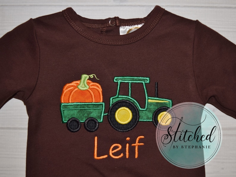 May include: Brown long-sleeve baby bodysuit with an embroidered design. The design features a green tractor pulling a wagon with an orange pumpkin. The name "Leif" is embroidered in orange. The text "Stitched by Stephanie" is in a circle.