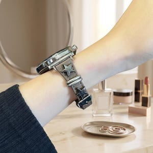 May include: A wristwatch featuring a dark gray leather strap adorned with metallic silver star-shaped studs and silver-toned hardware. The watch case is circular with a metallic finish, and the strap includes a matching silver-toned buckle closure.