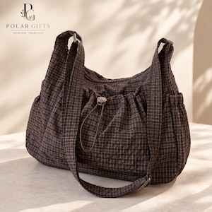 Puede incluir: Un bolso hobo de color marrón oscuro con un patrón de cuadrícula blanca, un bolsillo frontal fruncido con cierre de cordón ajustable y una correa para el hombro. El bolso tiene una textura acolchada suave y bolsillos laterales.