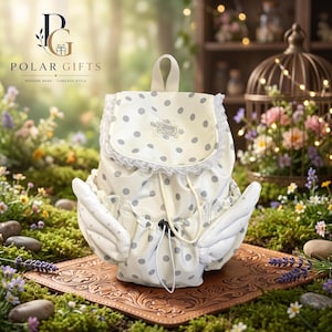 May include: A cream-colored backpack featuring a gray polka dot pattern, white lace trim along the flap, and two padded wing-shaped side pockets. The bag includes a drawstring closure with a black toggle and a small rectangular label on the front.