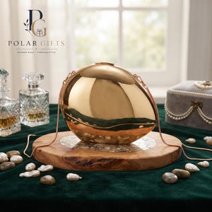 May include: A polished gold-tone egg-shaped clutch bag with a smooth, reflective surface and a thin matching gold-tone chain strap. The bag features a secure top clasp closure and a rounded, symmetrical design that mimics the shape of an egg.