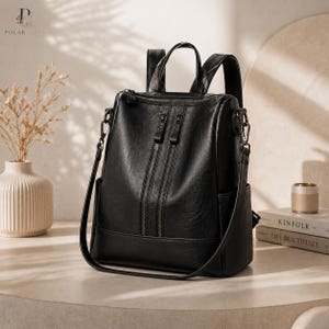 Pode incluir: Uma mochila preta com acabamento liso, apresentando dois bolsos verticais com zíper na frente, uma alça superior, alças de ombro ajustáveis e bolsos laterais. A bolsa inclui uma alça de ombro removível fixada em argolas de metal.