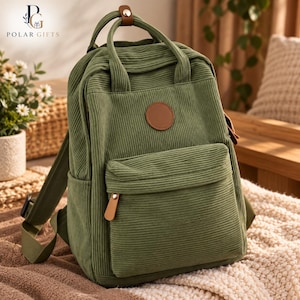 Op de afbeelding: Een olijfgroene rugzak van corduroy met een voorvak met ritssluiting, twee handvatten aan de bovenkant en een ronde bruine patch in lederlook. De tas is voorzien van verstelbare schouderbanden en bruine accenten op de ritstrekker.