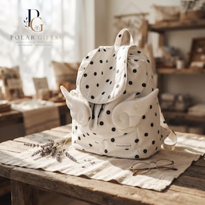 Puede incluir: Una mochila blanca con estampado de lunares negros y dos apliques de tela blanca acolchada en forma de alas en los laterales. La bolsa tiene una solapa superior, cierre de cordón y una pequeña etiqueta rectangular con el texto "nena boutique".