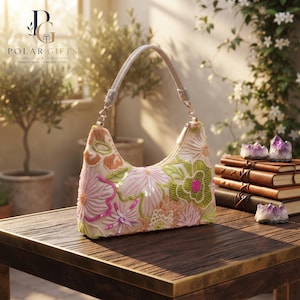 Pode incluir: Uma bolsa de mão tipo hobo na cor creme com bordados florais intrincados em tons de rosa, verde e laranja, acentuada com lantejoulas brilhantes. A bolsa possui uma alça metálica única e fecho de zíper, exibindo uma superfície texturizada.