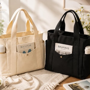 May include: Two canvas tote bags, one cream and one black, each featuring two top handles, side pockets holding white cylindrical containers, and a front pocket with a snap button closure. The cream bag holds a book labelled Matisse, and the black bag holds a book labelled Bauhaus.