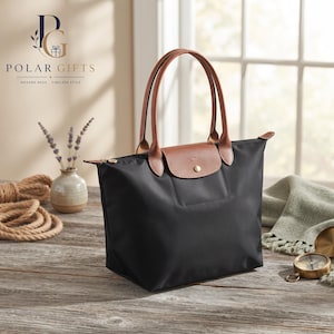 May include: A black nylon tote bag with brown leather handles and a brown leather flap closure featuring a gold-tone snap button. The bag has a structured rectangular base and a zipper closure along the top edge, displaying a clean, functional design.