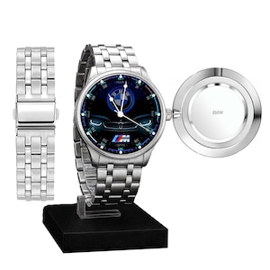 Pode incluir: Um relógio de pulso em aço inoxidável prateado com um mostrador redondo apresentando o logótipo azul escuro da BMW, um gráfico da frente de um carro e o logótipo M. O relógio tem uma bracelete de elos metálicos e uma tampa gravada BMW.